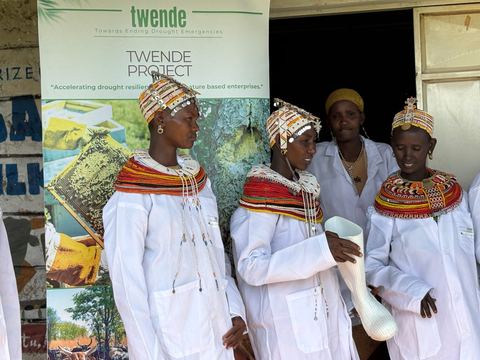 Women from a camel milk enterprise in white coats standing beside a TWENDE project banner